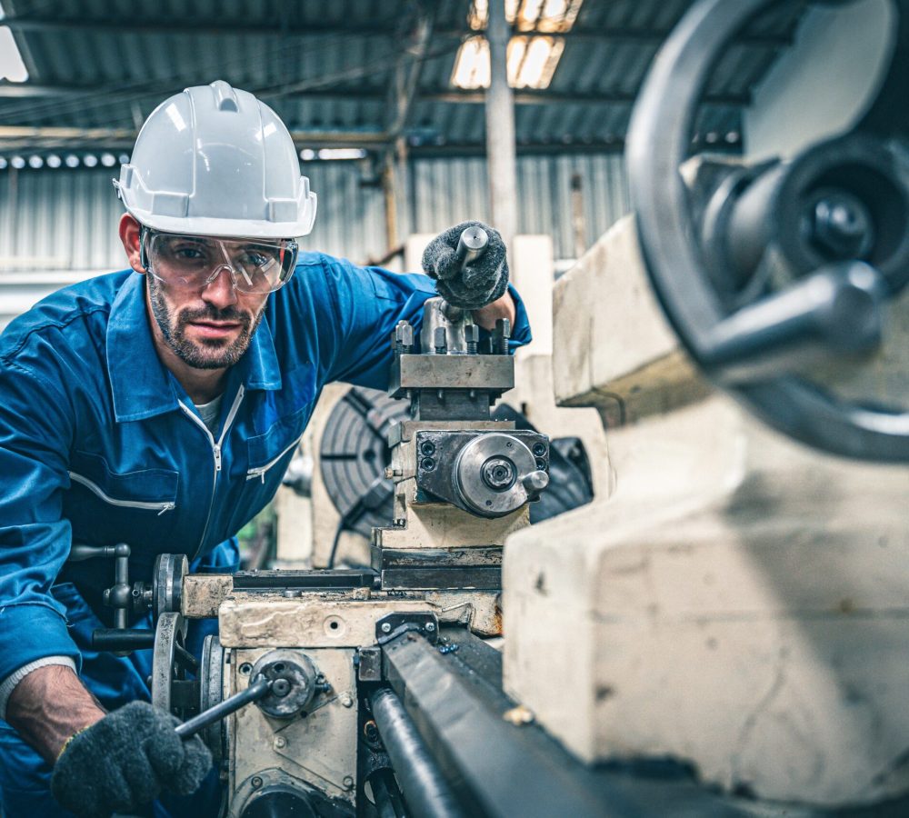 Male worker in blue jumpsuit and white hardhat operating lathe machine.